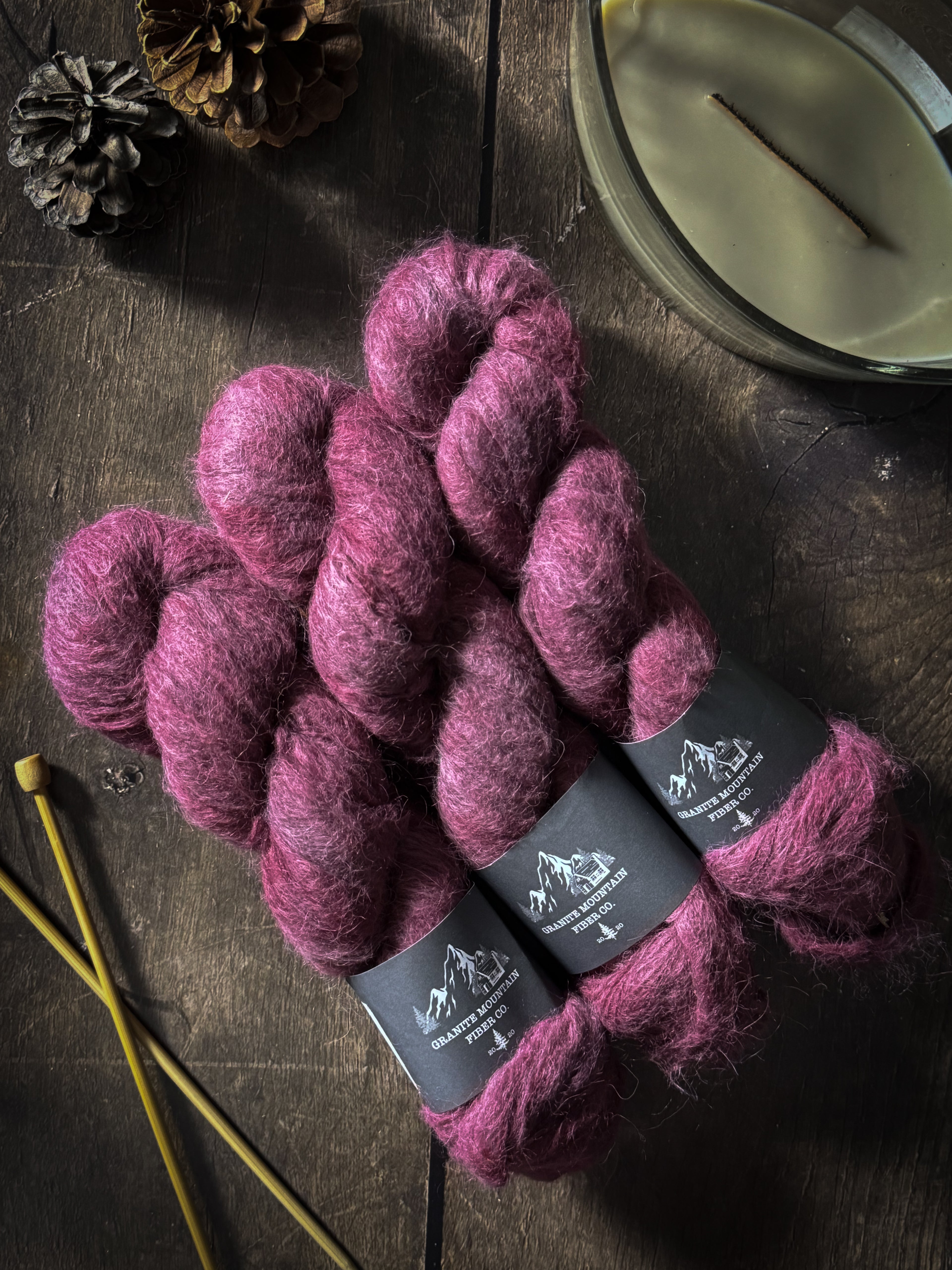 Three skeins of purple yarn with labels on a wooden surface, accompanied by knitting needles and a pine cone.